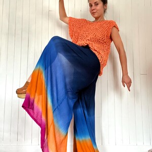 May include: An orange crochet top paired with wide-leg pants in a vibrant tie-dye pattern of blue, orange, and pink. The pants have a flowing, airy design. The outfit is worn with wooden clogs.