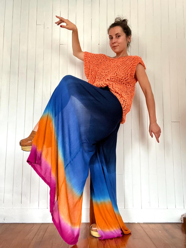 May include: An orange crochet top paired with wide-leg pants in a vibrant tie-dye pattern of blue, orange, and pink. The pants have a flowing, airy design. The outfit is worn with wooden clogs.