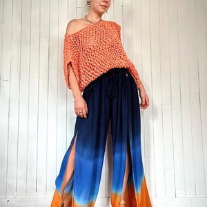 May include: An orange, open-weave, off-the-shoulder top paired with navy blue, split-leg pants. The pants feature a gradient of blue, orange, and pink. The outfit is styled with wooden platform sandals.