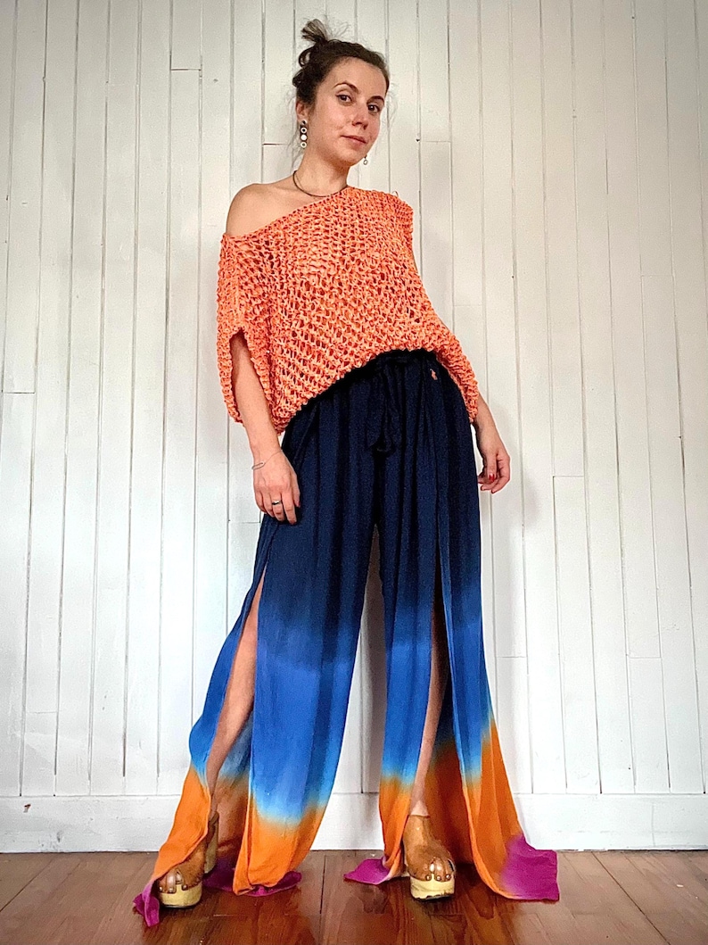May include: An orange, open-weave, off-the-shoulder top paired with navy blue, split-leg pants. The pants feature a gradient of blue, orange, and pink. The outfit is styled with wooden platform sandals.