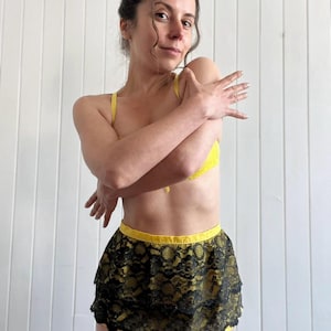 Op de afbeelding: Een vrouw draagt een gele bh en een zwarte kanten rok met een gele tailleband. De rok is gelaagd en heeft een geschulpte rand. De vrouw staat voor een witte muur.