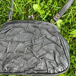 K&ouml;nnte beinhalten: Eine schwarze Clutch-Tasche mit silbernem Verschluss und d&uuml;nnem Schulterriemen. Die Tasche hat eine geraffte, faltige Textur. Der Hintergrund ist gr&uuml;nes Gras.