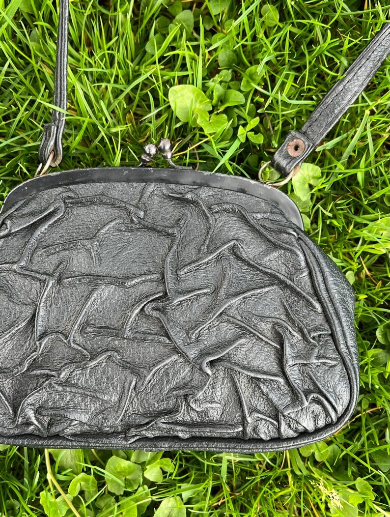 K&ouml;nnte beinhalten: Eine schwarze Clutch-Tasche mit silbernem Verschluss und d&uuml;nnem Schulterriemen. Die Tasche hat eine geraffte, faltige Textur. Der Hintergrund ist gr&uuml;nes Gras.