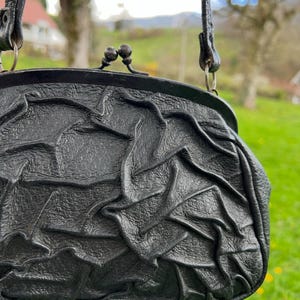 K&ouml;nnte beinhalten: Schwarze Lederhandtasche mit strukturiertem, ger&uuml;schtem Design. Die Tasche hat einen Verschluss und einen d&uuml;nnen Riemen. Der Hintergrund ist eine verschwommene Ansicht von gr&uuml;nem Gras und B&auml;umen.