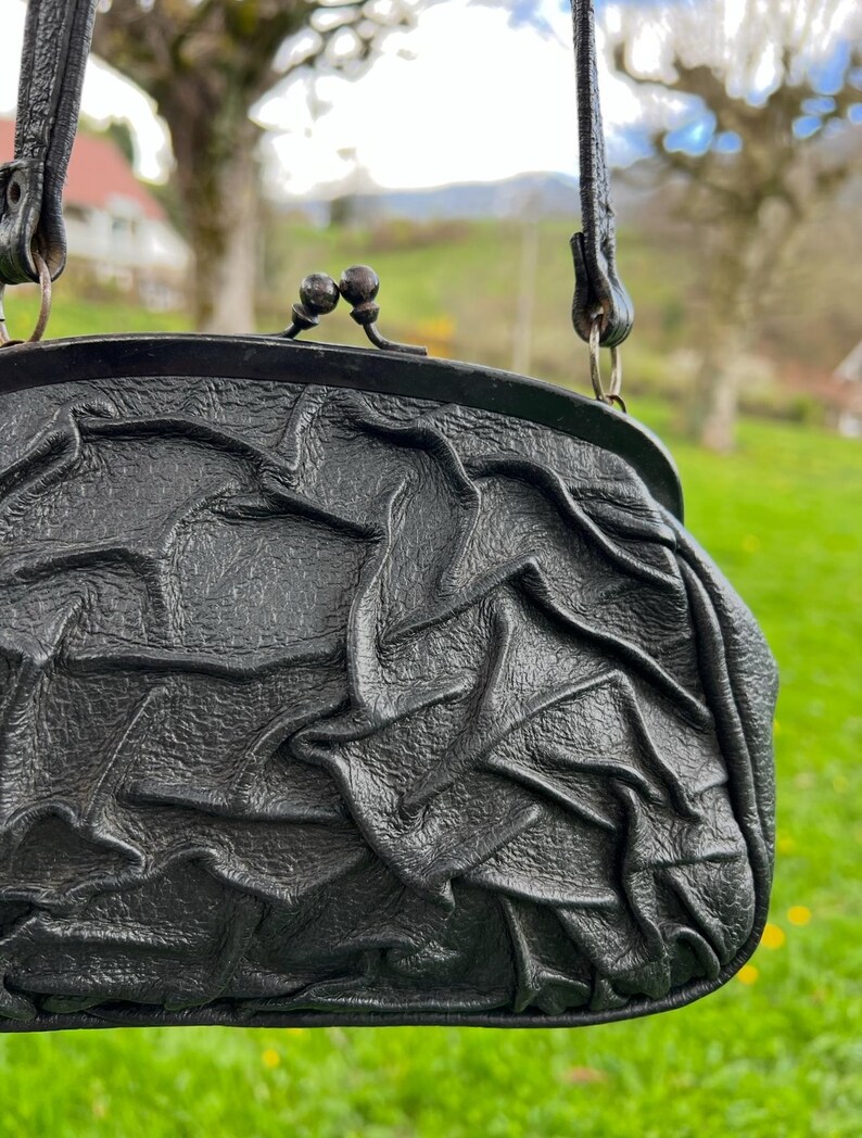 K&ouml;nnte beinhalten: Schwarze Lederhandtasche mit strukturiertem, ger&uuml;schtem Design. Die Tasche hat einen Verschluss und einen d&uuml;nnen Riemen. Der Hintergrund ist eine verschwommene Ansicht von gr&uuml;nem Gras und B&auml;umen.