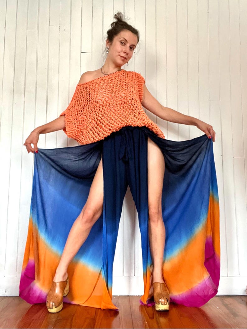 May include: An orange crochet top with a one-shoulder design is paired with navy blue pants featuring high slits and a colorful, tie-dye effect. The pants transition from blue to orange and pink. The model is wearing wooden clogs.