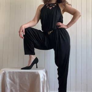 Könnte beinhalten: Ein schwarzer Jumpsuit mit einem Halterneck und einem goldenen Gürtel. Der Jumpsuit hat eine lockere Passform und ist aus einem weichen, fließenden Stoff gefertigt.
