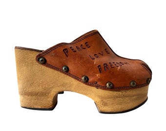 May include: Brown leather clog with a wooden platform sole. The leather upper has the words "Peace Love Freedom" embossed on it.