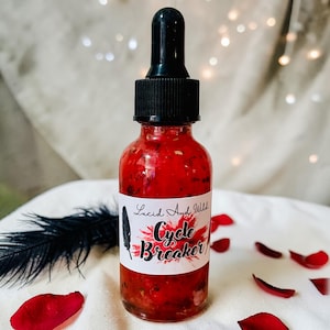 May include: A red glass bottle with a black dropper top. The bottle has a white label with the text "Lucid And Wild Cycle Breaker" and a feather illustration. The bottle is filled with a red liquid and surrounded by rose petals.