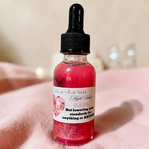 May include: A clear glass bottle with a black dropper top, filled with a pink liquid. The bottle has a white label with the text "Lucid And Wild High Value BABE" and a heart graphic. The liquid appears to have glitter.