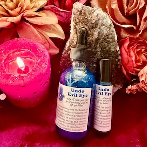 May include: A blue glass dropper bottle and a smaller blue glass roller bottle, both labeled Undo Evil Eye, featuring text about protection. The bottles are positioned in front of a large, jagged, light-colored crystal and pink flowers.