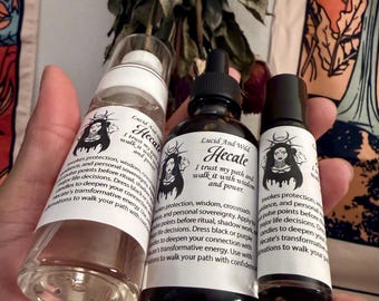 Puede incluir: Tres frascos de vidrio etiquetados como Hecate de Lucid And Wild. El frasco izquierdo tiene una bomba transparente, el central un gotero negro y el derecho una tapa de bola negra. Cada etiqueta blanca presenta una ilustración negra de una figura.