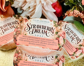 Könnte beinhalten: Drei runde Stück Strawberry Dream Seife in Klarsichtfolie verpackt. Jedes Etikett zeigt florale Grafiken, eine Kristallillustration und den Text: Wash with intention to invite sweetness, romance & loving energy. Enhance your allure while opening yourself to affection, pleasure & dreamy connections.