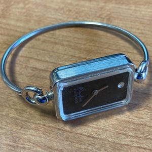 May include: A silver-toned bracelet watch with a rectangular black face. The watch face has a small diamond accent and the brand name "Crawford" is printed on the face. The watch band is a simple, curved design.