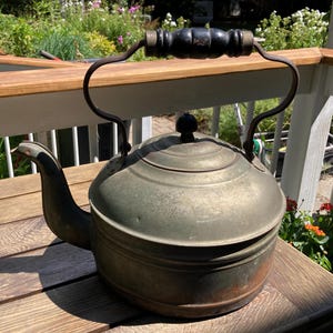May include: An antique metal kettle with a black wooden handle and a curved metal handle. The kettle has a spout and a lid with a small black knob. The kettle is sitting on a wooden surface.