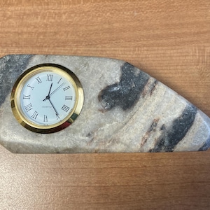 May include: A small quartz clock with a gold-colored bezel and white face, set into a uniquely shaped stone base. The stone has a mix of gray, black, and beige tones. The clock displays Roman numerals.