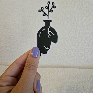 Puede incluir: Una pegatina en blanco y negro que presenta la silueta estilizada de una cabeza con forma de jarrón con una planta creciendo en la parte superior. El diseño incluye detalles de líneas blancas y el texto 1 COR. 4:17 impreso en el lateral.
