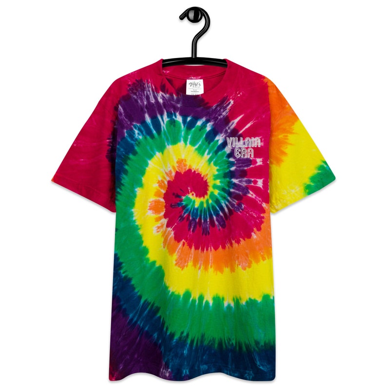 Puede incluir: Una camiseta tie-dye con un patr&oacute;n de espiral arco&iacute;ris y el texto "VILLAIN ERA" en letras blancas.