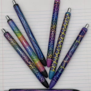 May include: A collection of seven glitter pens with various designs. The pens feature a range of colors, including rainbow, blue, purple, and yellow, with black tips and silver accents. Some pens have a leopard print pattern.