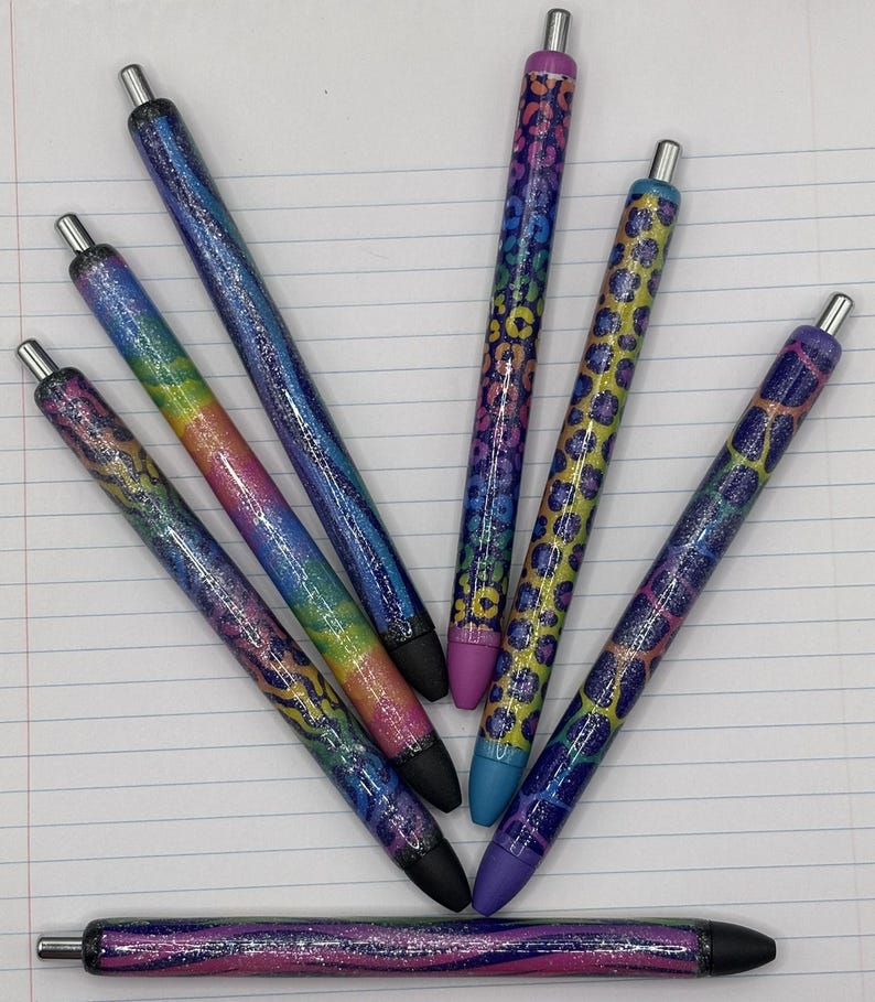 May include: A collection of seven glitter pens with various designs. The pens feature a range of colors, including rainbow, blue, purple, and yellow, with black tips and silver accents. Some pens have a leopard print pattern.