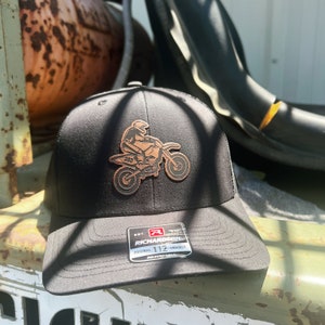 Op de afbeelding: Zwarte truckercap met een bruine leren patch met een motocrossrijder op een crossmotor. De patch heeft de tekst "365". De cap heeft een verstelbare band met de tekst "EST. 112 RICHARDSON ORIGINAL TRUCKER ADJUSTABLE".