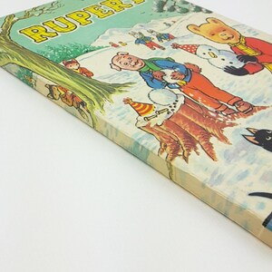 K&ouml;nnte beinhalten: Ein Vintage-Buchcover mit einer Cartoon-Illustration eines B&auml;ren, eines Schweins und anderer Figuren in einer verschneiten Winterlandschaft. Der Buchtitel lautet "Rupert The Daily Mirror"