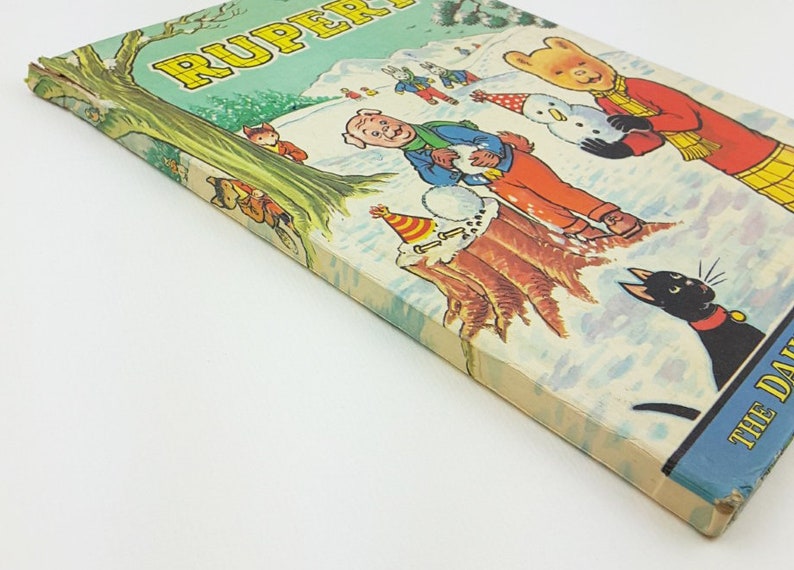 K&ouml;nnte beinhalten: Ein Vintage-Buchcover mit einer Cartoon-Illustration eines B&auml;ren, eines Schweins und anderer Figuren in einer verschneiten Winterlandschaft. Der Buchtitel lautet "Rupert The Daily Mirror"