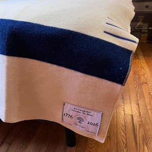 K&ouml;nnte beinhalten: Eine wei&szlig;e und blaue gestreifte Wolldecke mit einem braunen Etikett, auf dem "250th Commemorative Revolutionary War Blanket 1776 Liberty Tree 2026" steht.