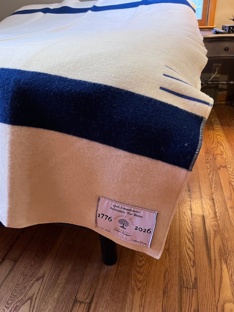 K&ouml;nnte beinhalten: Eine wei&szlig;e und blaue gestreifte Wolldecke mit einem braunen Etikett, auf dem "250th Commemorative Revolutionary War Blanket 1776 Liberty Tree 2026" steht.