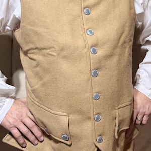 May include: A tan wool vest with silver buttons and two front pockets. The vest is part of a historical costume, paired with matching pants and a white collared shirt. The buttons are round and have a silver finish.