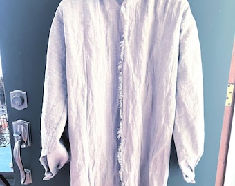 May include: A light blue linen shirt with long sleeves and a collar, hanging on a black hanger. The shirt has a button-up front with a frayed trim down the center and at the hem.