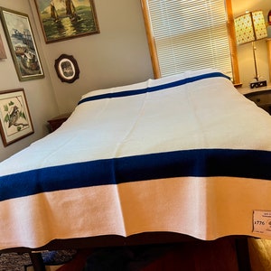 K&ouml;nnte beinhalten: Eine wei&szlig;e Wolldecke mit zwei dunkelblauen Streifen und einem pfirsichfarbenen unteren Rand. Die Decke hat ein Etikett mit dem Text "Old Commonwealth Blankets The Maine 1776 2026"