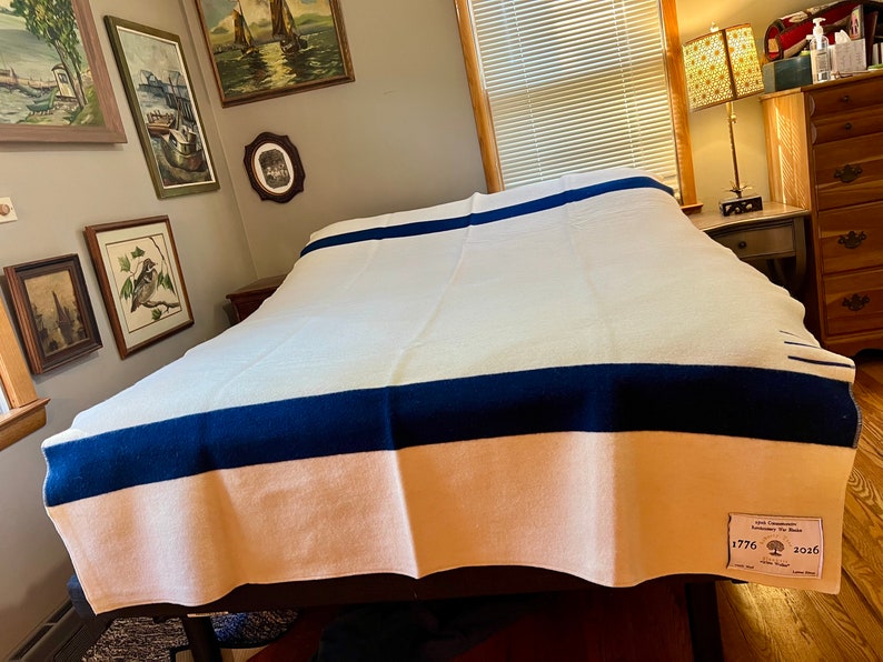 K&ouml;nnte beinhalten: Eine wei&szlig;e Wolldecke mit zwei dunkelblauen Streifen und einem pfirsichfarbenen unteren Rand. Die Decke hat ein Etikett mit dem Text "Old Commonwealth Blankets The Maine 1776 2026"