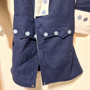 May include: Close-up of a navy blue wool coat with cream-colored accents. The coat features decorative silver buttons and pocket flaps. The coat is part of a historical costume or uniform.