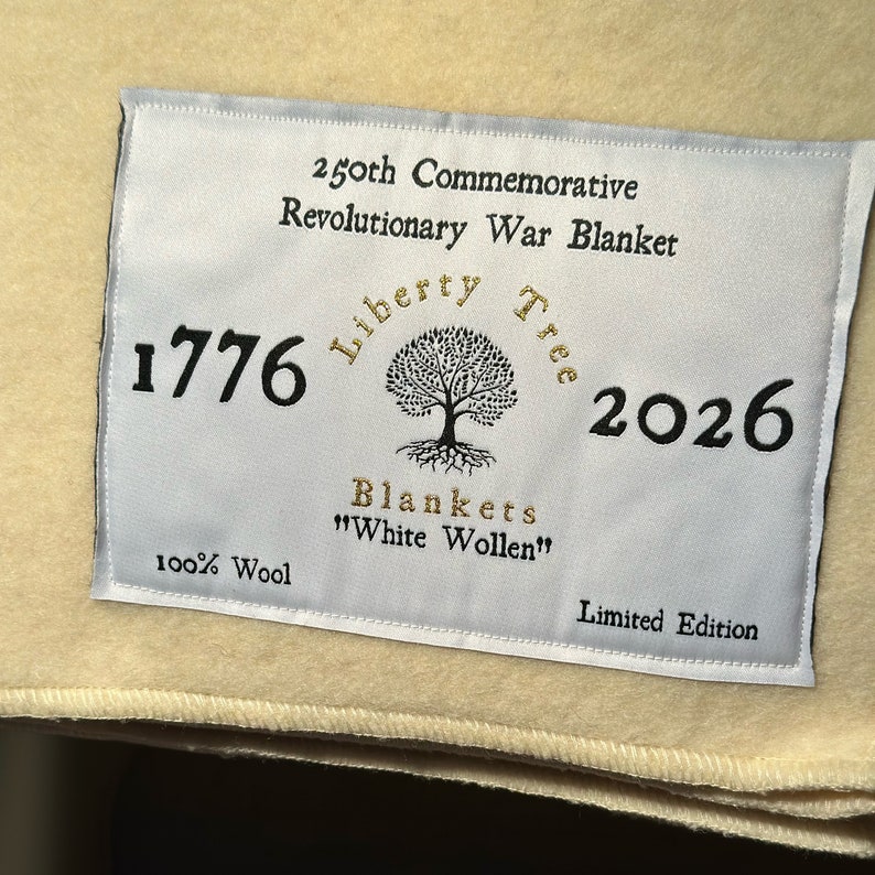 K&ouml;nnte beinhalten: Ein wei&szlig;es Etikett mit schwarzem Text, der "250th Commemorative Revolutionary War Blanket 1776 Liberty Tree 2026 Blankets "White Wollen" 100% Wool Limited Edition" lautet. Das Etikett hat einen goldenen Baum mit Wurzeln und einen goldenen Kreis darum mit den Worten "Liberty Tree" in Gold.
