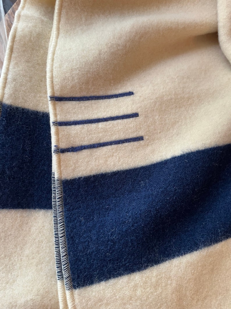 K&ouml;nnte beinhalten: Eine wei&szlig;-marineblaue gestreifte Decke mit einer gen&auml;hten Naht entlang der Kante. Die Decke hat drei horizontale Linien, die in Marineblau auf dem wei&szlig;en Abschnitt gen&auml;ht sind.