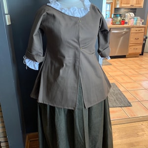 May include: A brown, long-sleeved jacket with a white ruffle collar and a green skirt. The jacket has a front closure with buttons and a slightly flared hem.