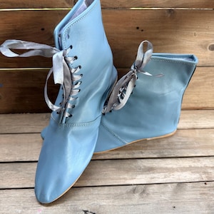 May include: A pair of light blue ankle boots with silver satin ribbon laces. The boots have a pointed toe and a low heel. The boots are made of a soft, leather-like material and are displayed on a wooden surface.