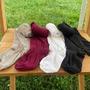Può includere: Quattro paia di calzini di diversi colori: beige, bordeaux, bianco e nero. I calzini sono arrotolati e posizionati su una superficie di legno.