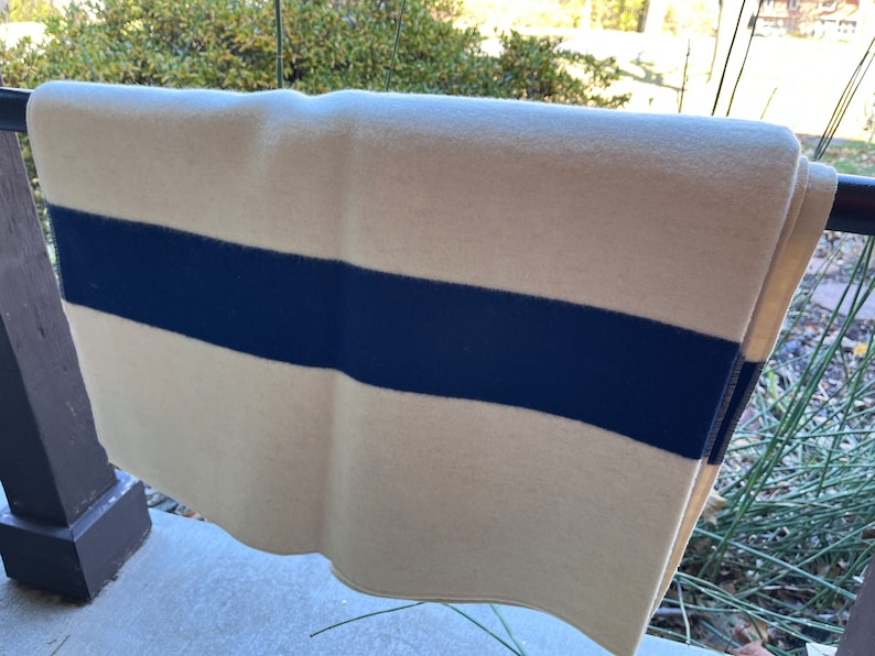 K&ouml;nnte beinhalten: Eine wei&szlig;e Wolldecke mit einem einzigen breiten dunkelblauen Streifen. Die Decke ist gefaltet und h&auml;ngt an einem Gel&auml;nder.