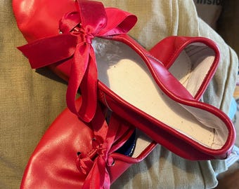 May include: A pair of bright red ballet flats with matching satin ribbons. The shoes have a rounded toe and a soft, white interior. The ribbons are tied in bows at the top of each shoe, adding a decorative touch. The shoes are designed for dance.