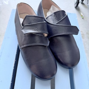 May include: A pair of dark navy blue leather shoes featuring a rounded toe design and a single wide strap across the top that secures with a hook-and-loop closure. The interior lining is a light beige color, and the shoes have a smooth finish.