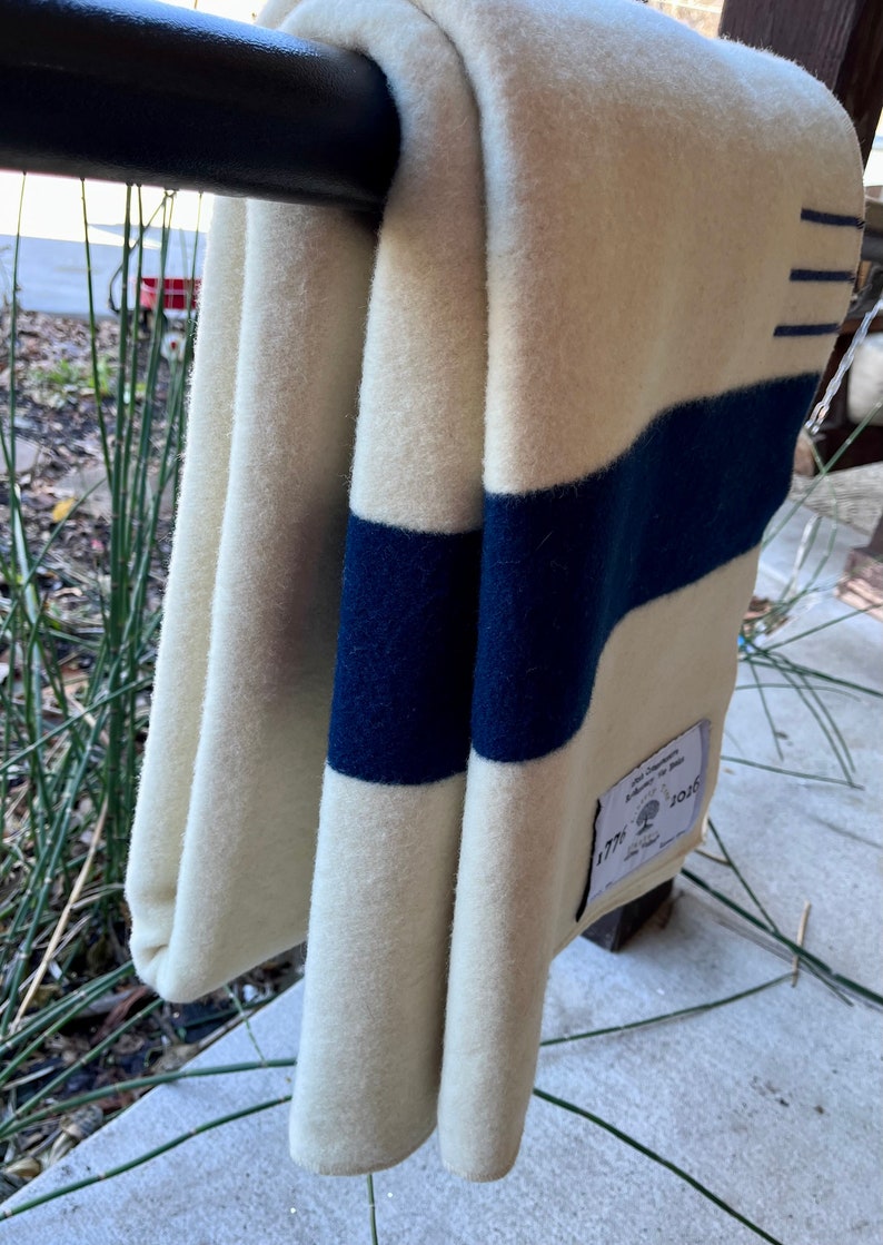 K&ouml;nnte beinhalten: Eine wei&szlig;e Wolldecke mit drei dunkelblauen Streifen. Die Decke ist gefaltet und h&auml;ngt an einem schwarzen Metallgel&auml;nder. Ein wei&szlig;es Etikett mit dem Text "1776" und einem Baumlogo ist an der Decke befestigt.