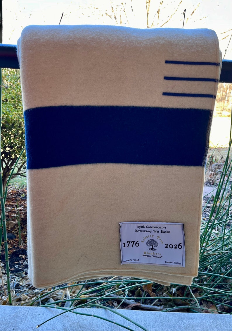 K&ouml;nnte beinhalten: Eine wei&szlig;e Wolldecke mit einem blauen Streifen und einem Etikett mit der Aufschrift "250. Jahrestag der Amerikanischen Revolution 1776 Liberty Tree Blankets "White Woolen" 2026 Limited Edition".