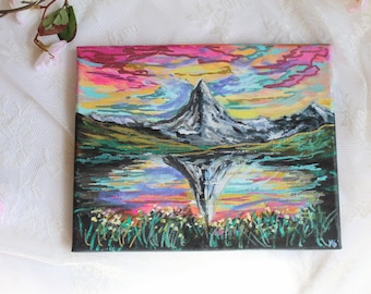 May include: A colorful painting of a mountain reflected in a lake. The sky is a vibrant mix of pink, yellow, and blue. The mountain is gray and white, and the lake is a dark blue. The painting is framed in black.