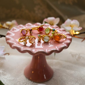 Könnte beinhalten: Eine Sammlung bunter Ringe mit Glassteinen und goldfarbenem Draht, der um sie herumgewickelt ist, auf einem rosafarbenen Kuchenständer ausgestellt.