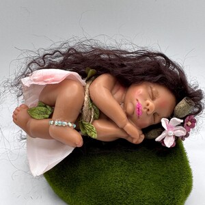 May include: A sleeping baby doll with dark hair, wearing a pink skirt and a bracelet. The doll is adorned with floral accents and rests on a green mossy surface. The doll's lips are painted pink.