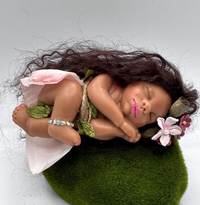 May include: A sleeping baby doll with dark hair, wearing a pink skirt and a bracelet. The doll is adorned with floral accents and rests on a green mossy surface. The doll's lips are painted pink.