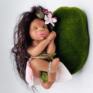 May include: A handcrafted baby doll with dark hair, adorned with flowers, and a pink lip color. The doll is nestled against a heart-shaped mossy green backdrop, with a light pink fabric and a beaded bracelet.