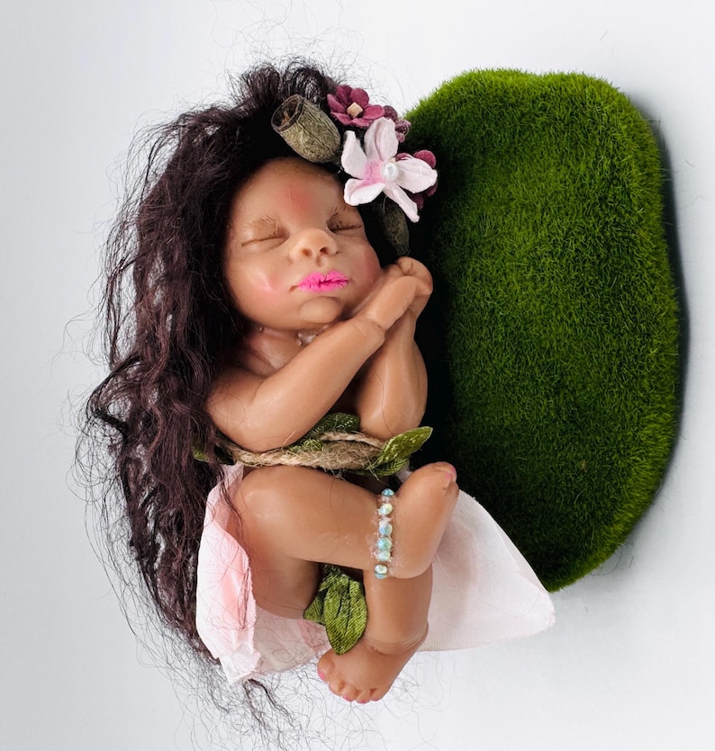 May include: A handcrafted baby doll with dark hair, adorned with flowers, and a pink lip color. The doll is nestled against a heart-shaped mossy green backdrop, with a light pink fabric and a beaded bracelet.